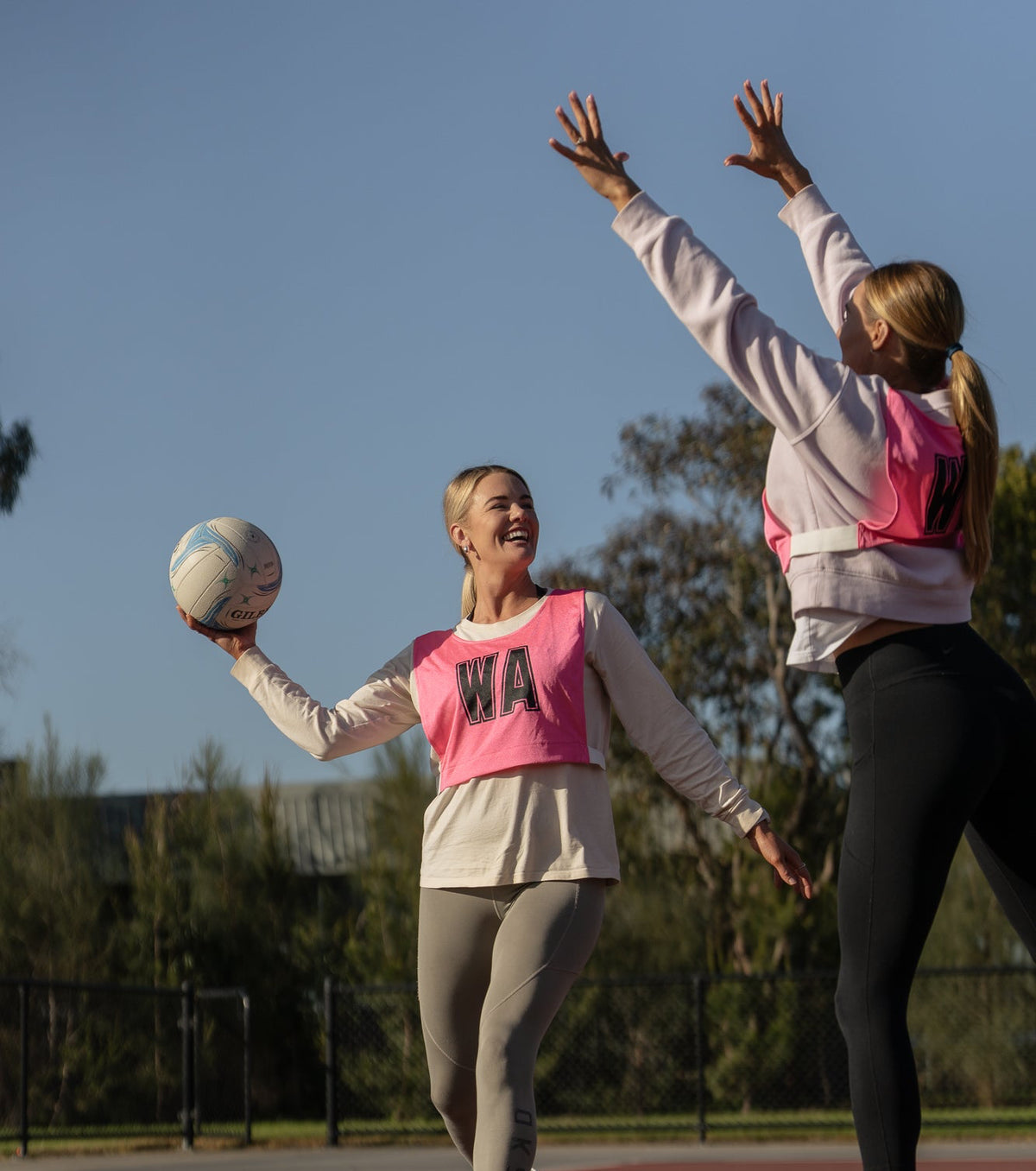 Two women playing netball on an outdoor court, wearing matching pink and white athletic vests over long-sleeve tops and leggings.