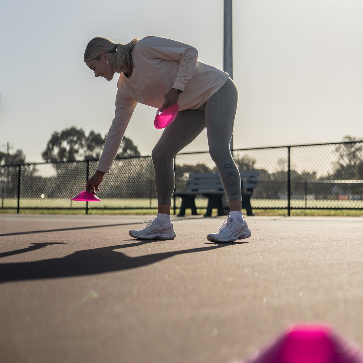 A woman in athletic wear bends down on an outdoor court while holding a bright pink disc, with another pink disc blurred in the foreground and a fence in the background.